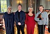 Fire voksne personer står samlet i et trepanelt rom med piano og instrumenter.