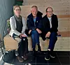 Tre personer sitter på en benk i et rom med trevegger, med en veske på gulvet.
