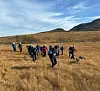Ei gruppe turgåere og to hunder går over et gyllent høyfjellsområde under blå himmel.