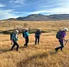 Fire fjellvandrere går gjennom gyllent terreng foran lave fjell.