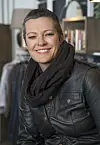 Smilende kvinne i svart skinnjakke og skjerf foran en bokhylle.