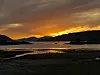 Solnedgang over en fjord med fjell, strand og speilinger i vannet.
