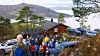 Turgruppe foran en trehytte i fjellet, med biler, furutrær og vann i bakgrunnen.