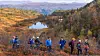 Turgåere i fargerikt høstlandskap foran et lite fjellvann og skogkledde fjell.