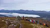Fotturister går langs en steinete fjellrygg med skyer og fjellandskap i bakgrunnen.