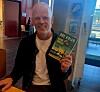 Smilende mann holder en grønn bok foran seg i et lyst interiør.