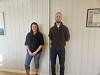 To voksne personer står innendørs foran en lys panelvegg med innrammede bilder.