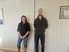 To voksne personer står innendørs foran en lys panelvegg med innrammede bilder.