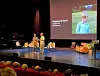 To personer presenterer på en scene foran et publikum, med et bilde av et barn på storskjerm.