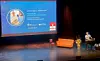 Foredragsholder ved talerstol foran en stor presentasjonsskjerm på en mørklagt scene.