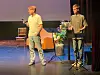To personer på en scene med mikrofoner, bord og blomster.