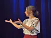 Kvinne med mønstret bluse gestikulerer foran mørkeblå scenetepper.