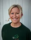 Smilende blond kvinne i mørkegrønn genser foran en lys vegg.