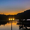 Tre lamper lyser ved en innsjø med speilblankt vann, mørke åser og brygge i skumringen.