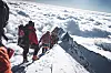Fjellklatrere i røde jakker går på en snødekt fjellrygg over skyene.