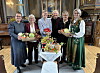 Ei gruppe voksne damer holder kurver med frukt, grønnsaker og blomster innendørs.