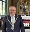 Smilende mann i mørk dress med kjede foran et utsmykket kirkealter.