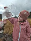 Barn i rosa klær holder en gjennomsiktig plastpose med vann og en liten fisk i, i et overskyet fjellandskap.