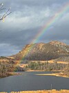Regnbue over skogkledde fjell og en innsjø under mørke skyer.