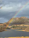 Regnbue over skogkledde fjell og en innsjø under mørke skyer.