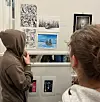 To besøkende ser på tegninger og grafiske kunstverk i et galleri.