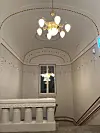 Utsmykket trappehall med lysekrone, buet tak, vindu og dekorative rekkverk.