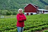 Kvinne i rosa jakke står foran en grønn åker og rød låve.