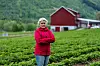 Kvinne i rosa jakke står foran en grønn åker og rød låve.