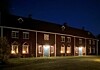 Rødbrun trebygning med lysende vinduer og dører mot en plen om kvelden.