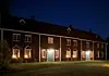 Rødbrun trebygning med lysende vinduer og dører mot en plen om kvelden.