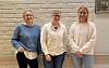 Tre kvinner i hverdagsklær står på rekke foran en lys murvegg.
