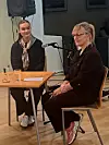 To kvinner sitter ved et bord med mikrofoner i et innendørs lokale.