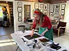 Kunstner arbeider ved et bord med bilder og utstyr i et atelier.