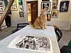 Rød katt sitter på et bord med et stort svart-hvitt trykk, omgitt av innrammet kunst.