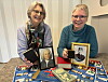To smilende kvinner holder innrammede portretter ved et bord med fargerikt materiale.
