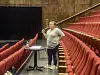 Person ved et bord i en teatersal med røde seter og svart sceneteppe.