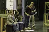 Gruppe personer i et teatermiljø, med noen sittende og andre stående ved siden av scenerekvisitter.