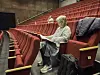 Tilskuer sitter i en rød teatersal med veske og jakke på setet ved siden av seg.