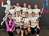 Ungt håndballag poserer i hvite og lilla drakter foran en sponsorvegg.