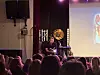 En foredragsholder sitter på scenen foran et publikum, med en projeksjon på lerretet til høyre.