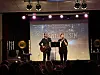 To personer står på en opplyst scene foran en projektorskjerm, med publikum i forgrunnen.