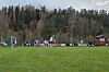 Fotballspillere i aksjon på en gressbane med skog og klubbhus i bakgrunnen.