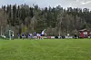 Fotballspillere i aksjon på en gressbane med skog og klubbhus i bakgrunnen.