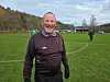 Smilende mann i mørkt treningstøy på en fotballbane med skog i bakgrunnen.