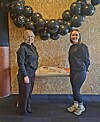 To kvinner poserer under en bue av svarte ballonger i et innendørs lokale.