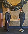 To kvinner poserer under en bue av svarte ballonger i et innendørs lokale.