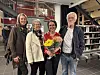 Fire personer står sammen innendørs; personen i midten holder en fargerik blomsterbukett.