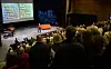Publikum ser mot en scene med en person, en opplyst skjerm og en oransje benk.