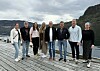 Åtte personer poserer på en trebrygge med fjord og fjell i bakgrunnen.