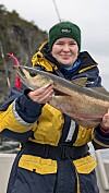 Jente i gult og mørkt regntøy holder en stor fisk ved en steinete kyst.
